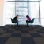 Two people sit facing each other in chairs by large windows in a bright lobby.