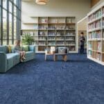 Bright library reading room with blue carpet, bookshelves, tables, and a person sitting by the window