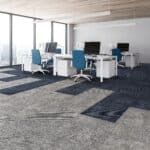 Modern office with desks, computer monitors, blue chairs, and large windows over gray carpet.