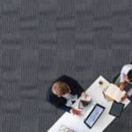 Overhead view of two people at a white table with a laptop, papers, and coffee cups on a gray floor
