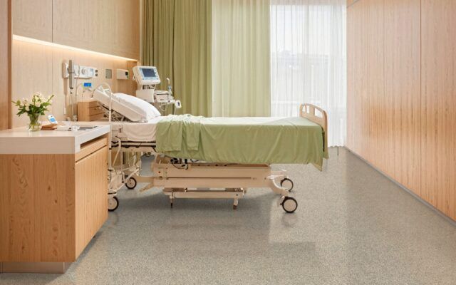 Hospital room with an empty bed, green blanket, and medical equipment beside a window.