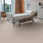 Hospital bed in a bright room beside a window and curtain, with medical equipment nearby.