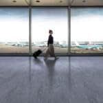 Traveler walking with suitcase through airport terminal by large windows and runway view