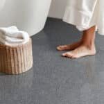Bare feet beside a white bed, with a woven basket or stool on a gray carpet.