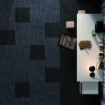 Top-down view of a white desk with a laptop, coffee cup, notebook, and small office items on dark carpet.