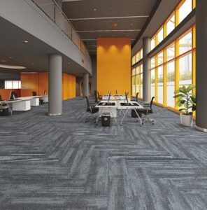 Modern office lounge with gray carpet, orange accent wall, tables, chairs, and tall windows bringing in daylight