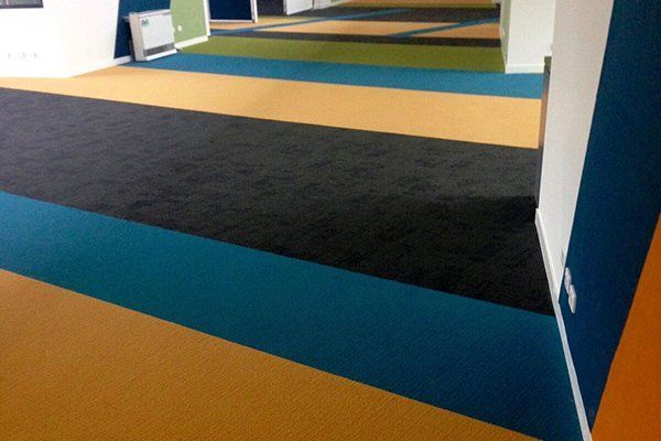 A wide hallway featuring floor carpet with alternating diagonal stripes of yellow, teal, black, and green.