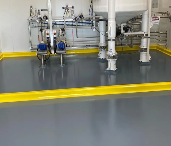 Industrial room with white pipes and blue pumps on a gray floor with yellow safety borders.