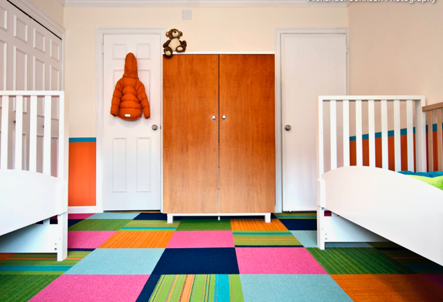 Colorful nursery with two white cribs, orange wardrobe, and multicolored geometric carpet