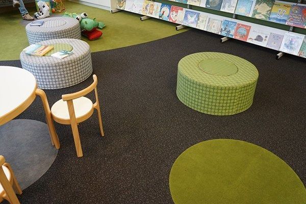 A children’s library area with round ottomans, a table with chairs, and wall-mounted shelves displaying children's books.