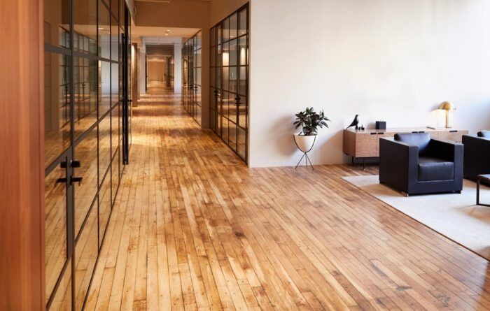 A bright office hallway with polished wood floors, glass-paneled walls, and a small sitting area with a black armchair.