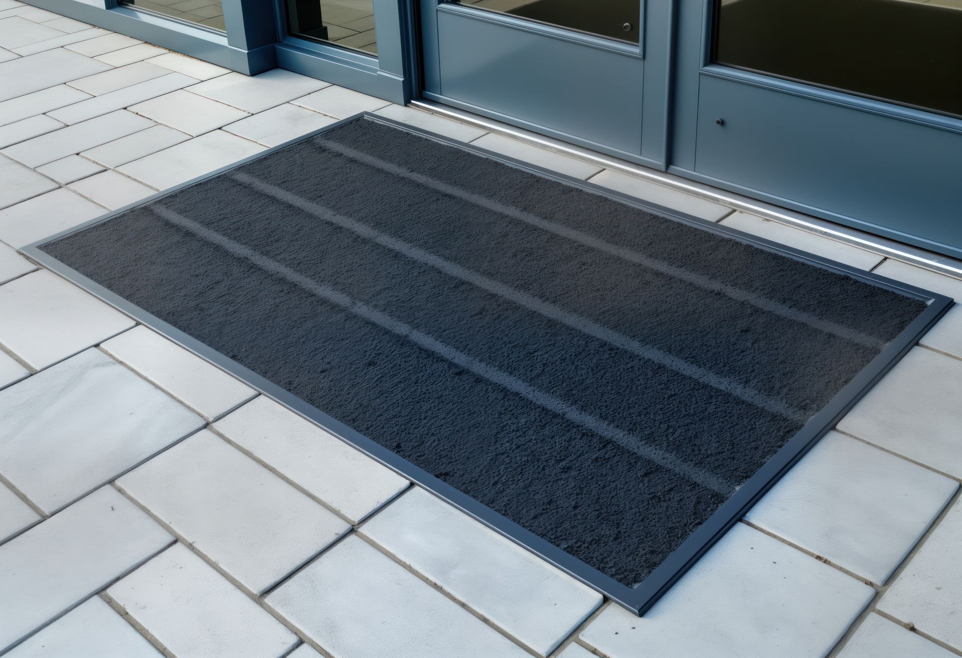 A dark gray, rectangular outdoor entrance mat with textured horizontal stripes placed on a light tile patio by glass doors.