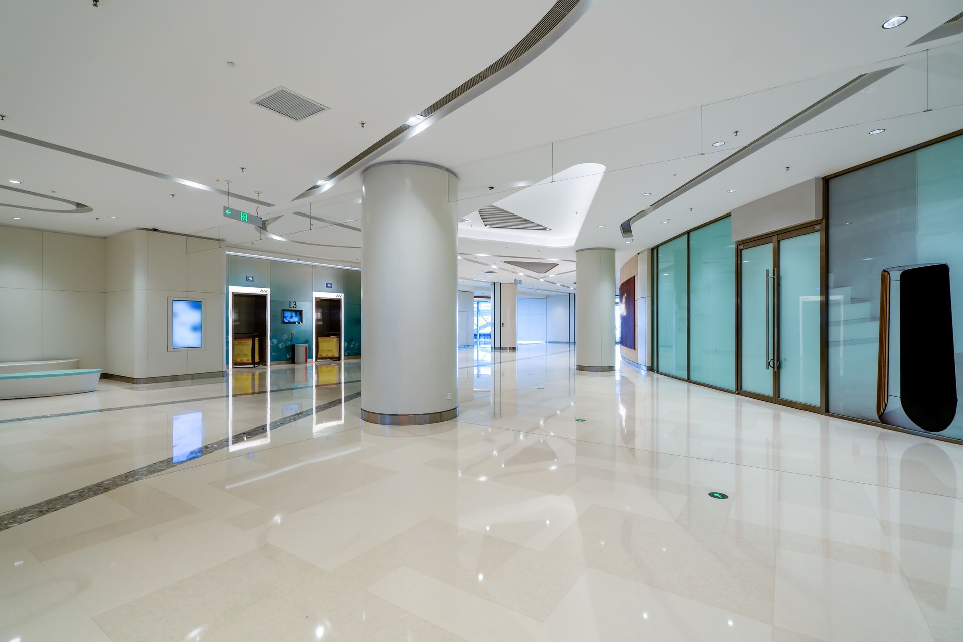 A spacious, brightly lit modern lobby with polished white floors, large support columns, and closed glass-fronted stores.