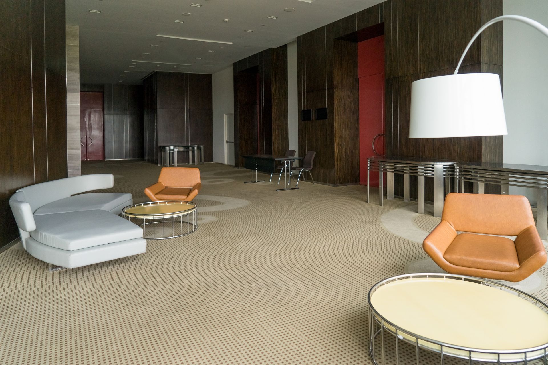 A modern, minimalist office lounge with a light gray curved sofa, two orange armchairs, coffee tables, and a floor lamp.