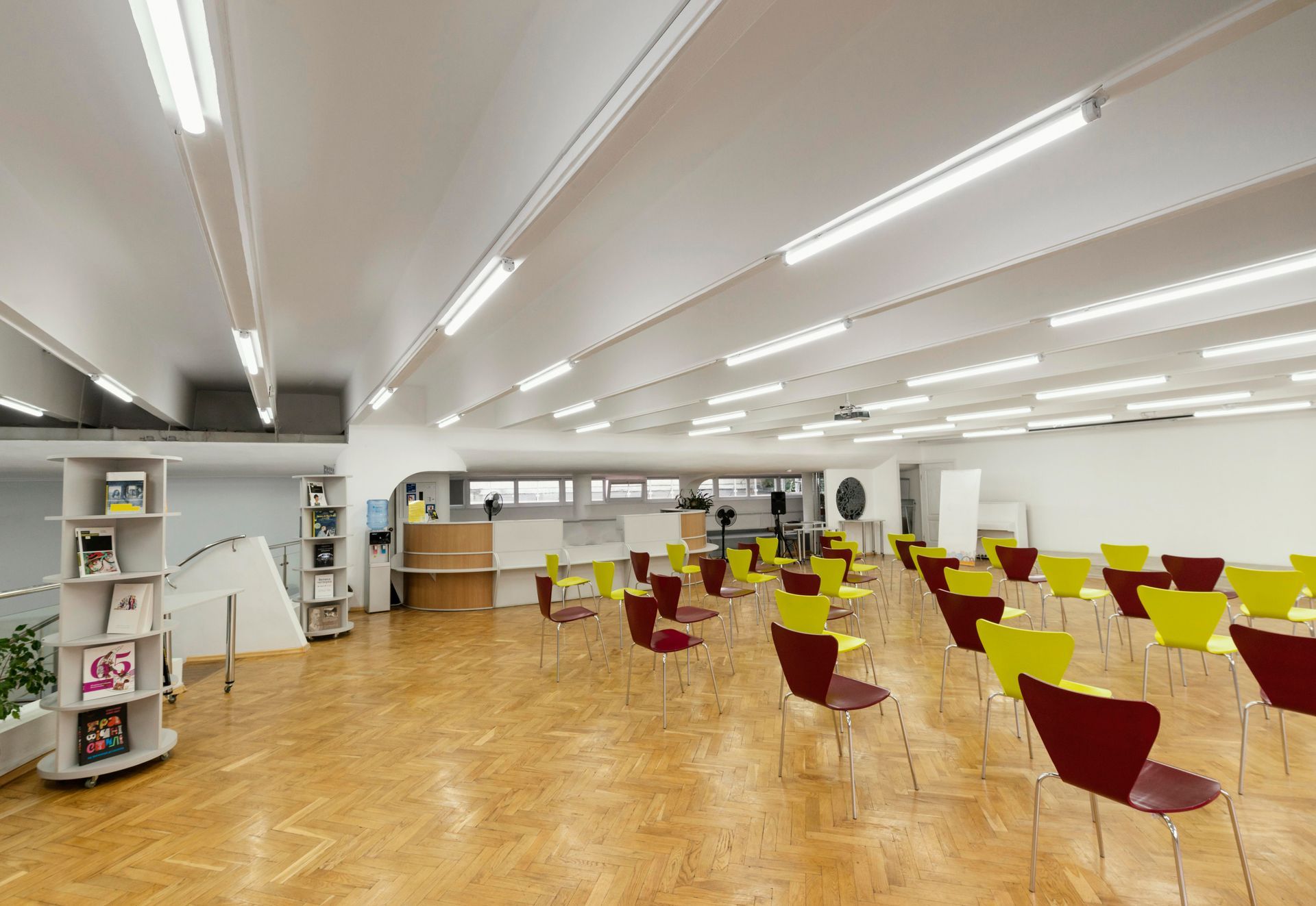 An empty conference room with light wood floors, fluorescent lighting, and rows of red and yellow chairs facing a desk.