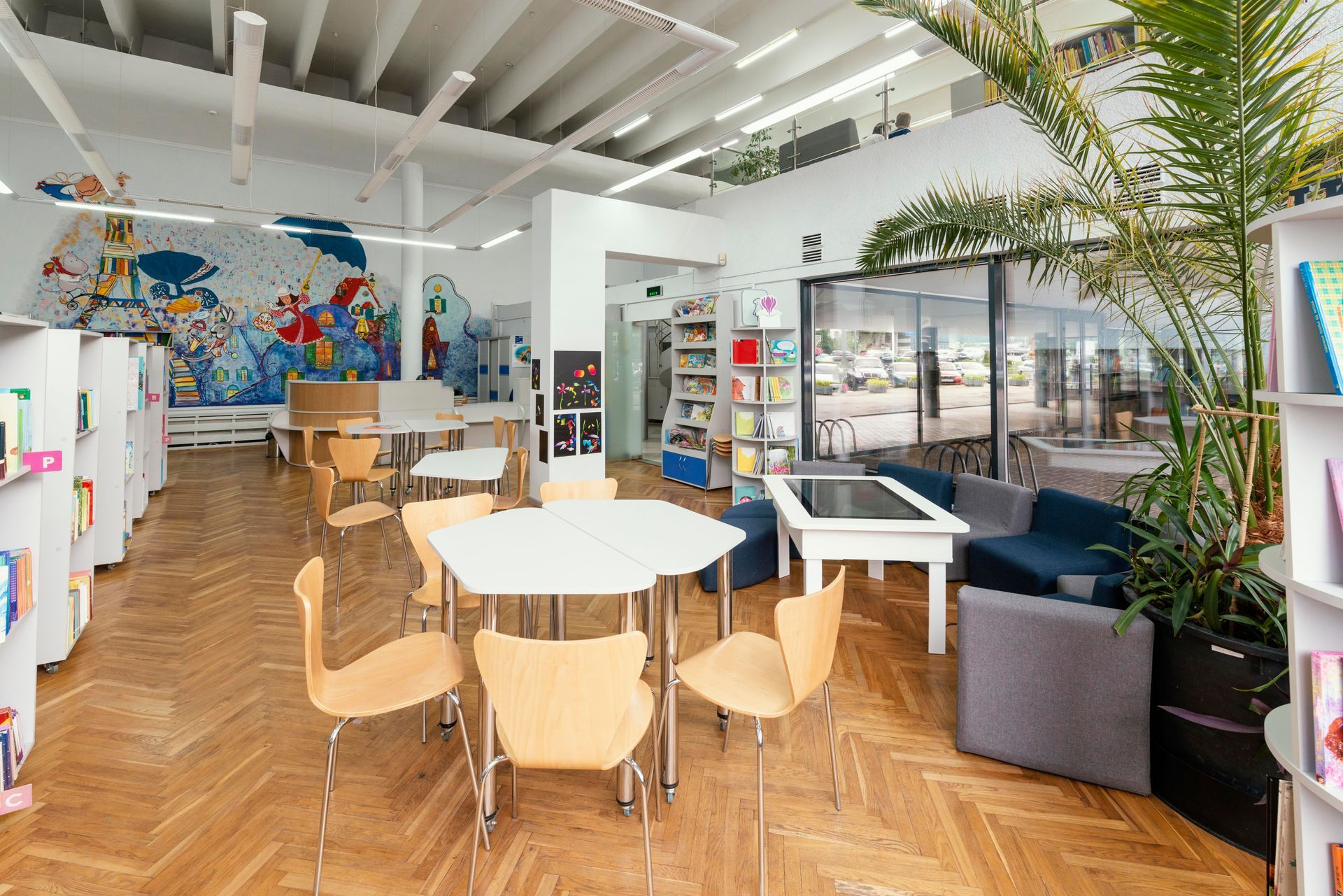 A modern library interior featuring hexagonal tables, chairs, bookshelves, a large wall mural, and a tall potted plant.