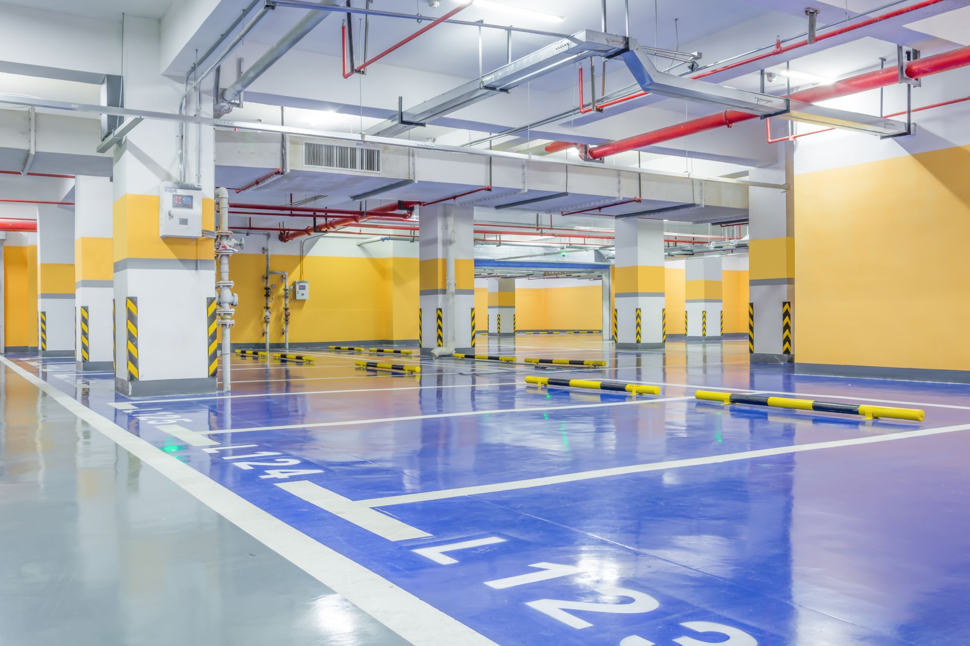 Empty underground parking garage with blue floor markings, yellow pillars, and overhead pipes
