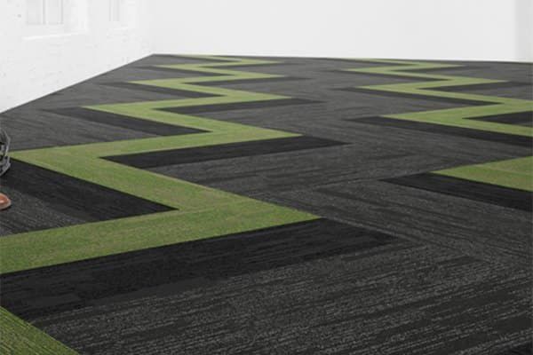 Modern office flooring featuring repeating lime green zigzag patterns across a textured dark gray carpet tile surface.