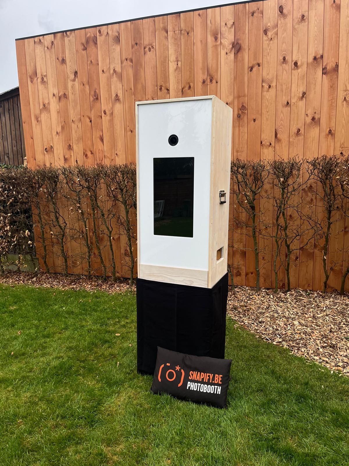Een houten photobooth staat op een zwarte sokkel op een gazon voor een houten hek, met een reclamebord ernaast.