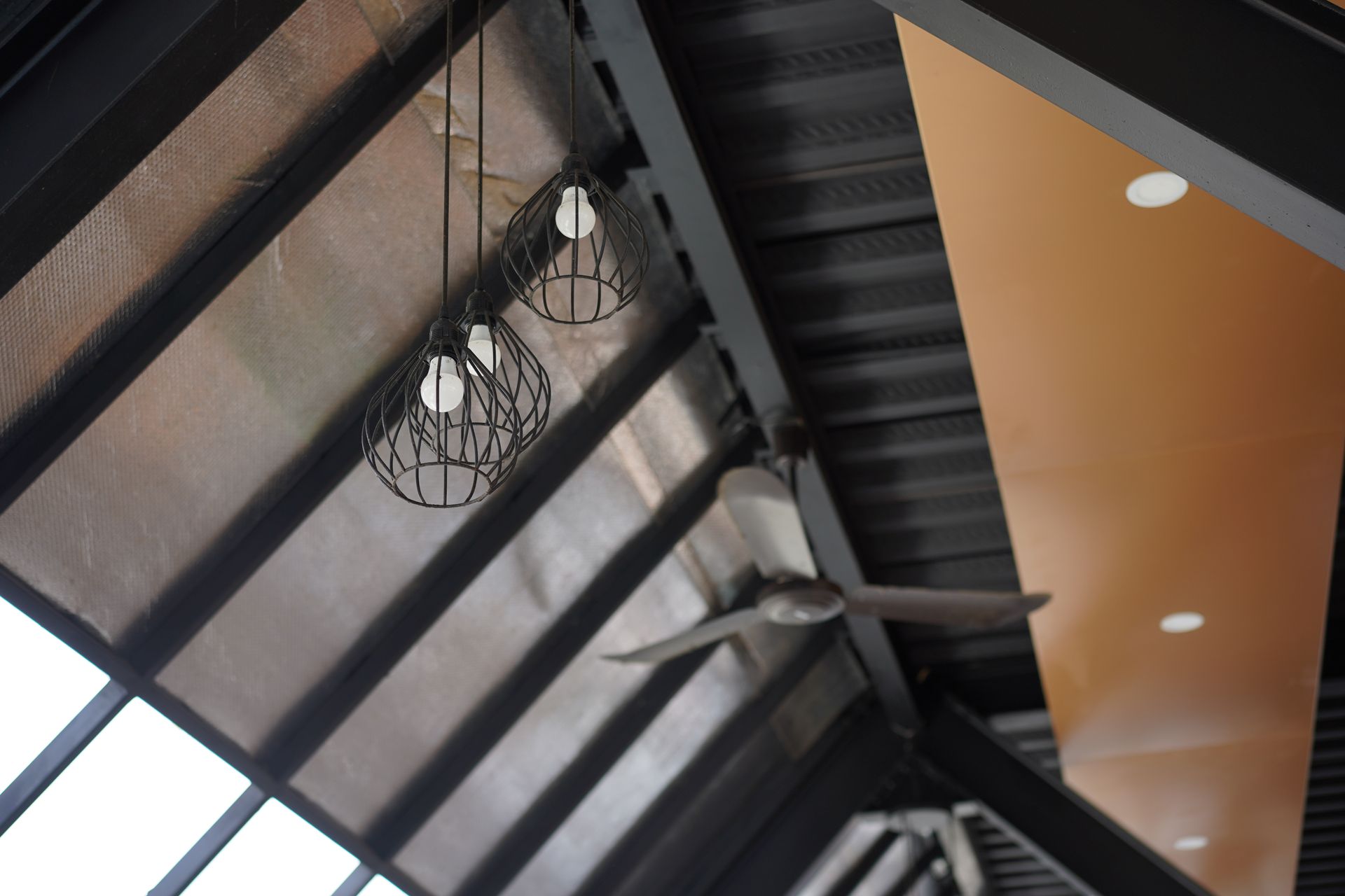 Angled view of a modern ceiling with black beams, hanging pendant lights, and a tan panel with recessed lights