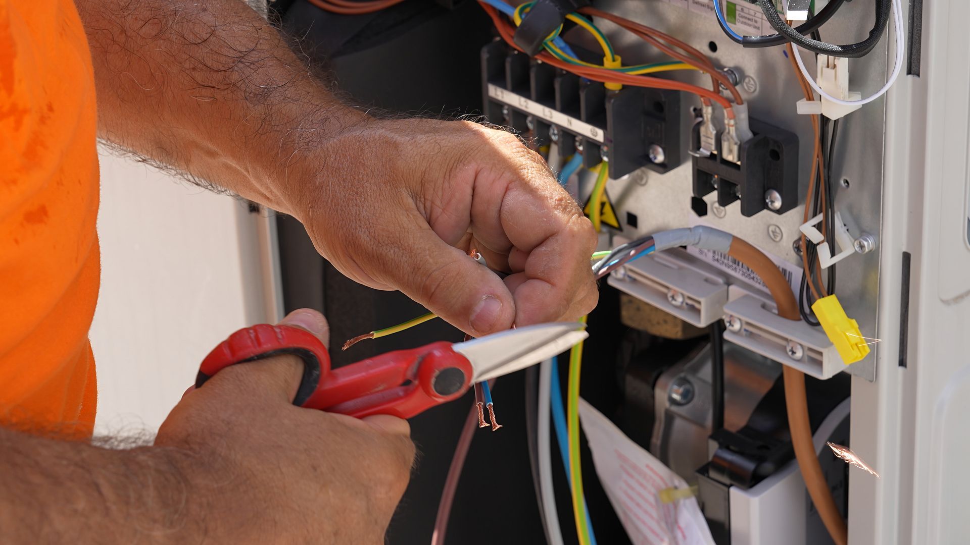 Electrician wiring a wall panel, using red pliers to cut a white cable beside exposed wires.
