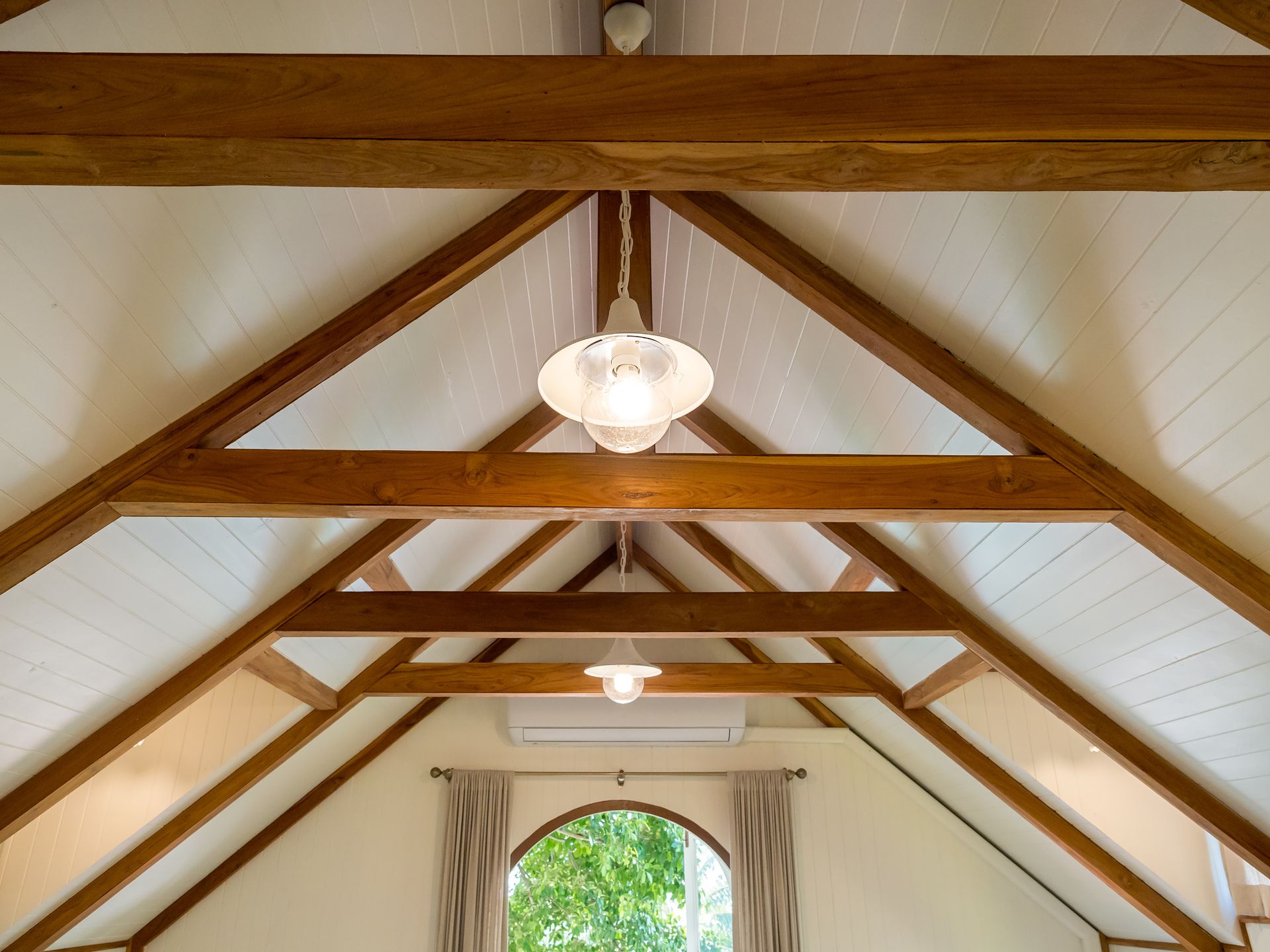 Wood-beamed vaulted ceiling with hanging lights and a window framed by curtains above greenery.