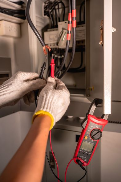 Gloved hands using a red multimeter to test wires inside an electrical panel