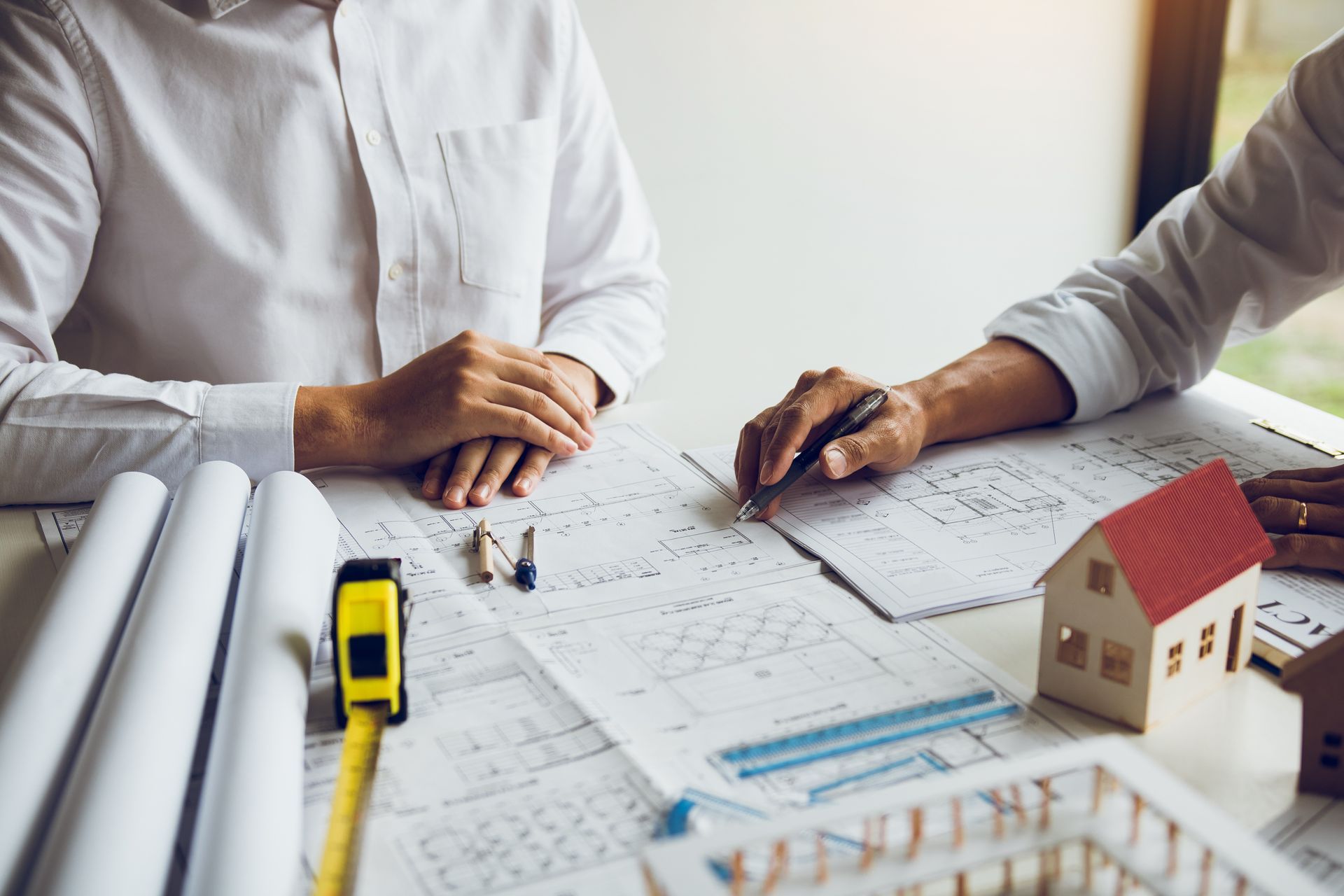 Two people reviewing architectural blueprints with a yellow tape measure and house model on a desk