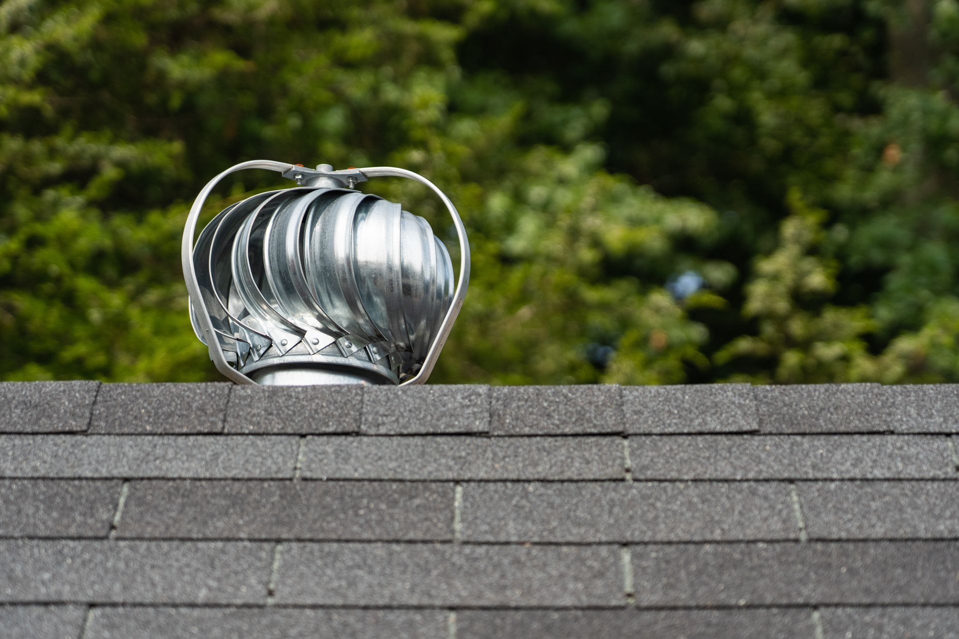 Silver turbine vent on a gray shingled roof with trees in the background
