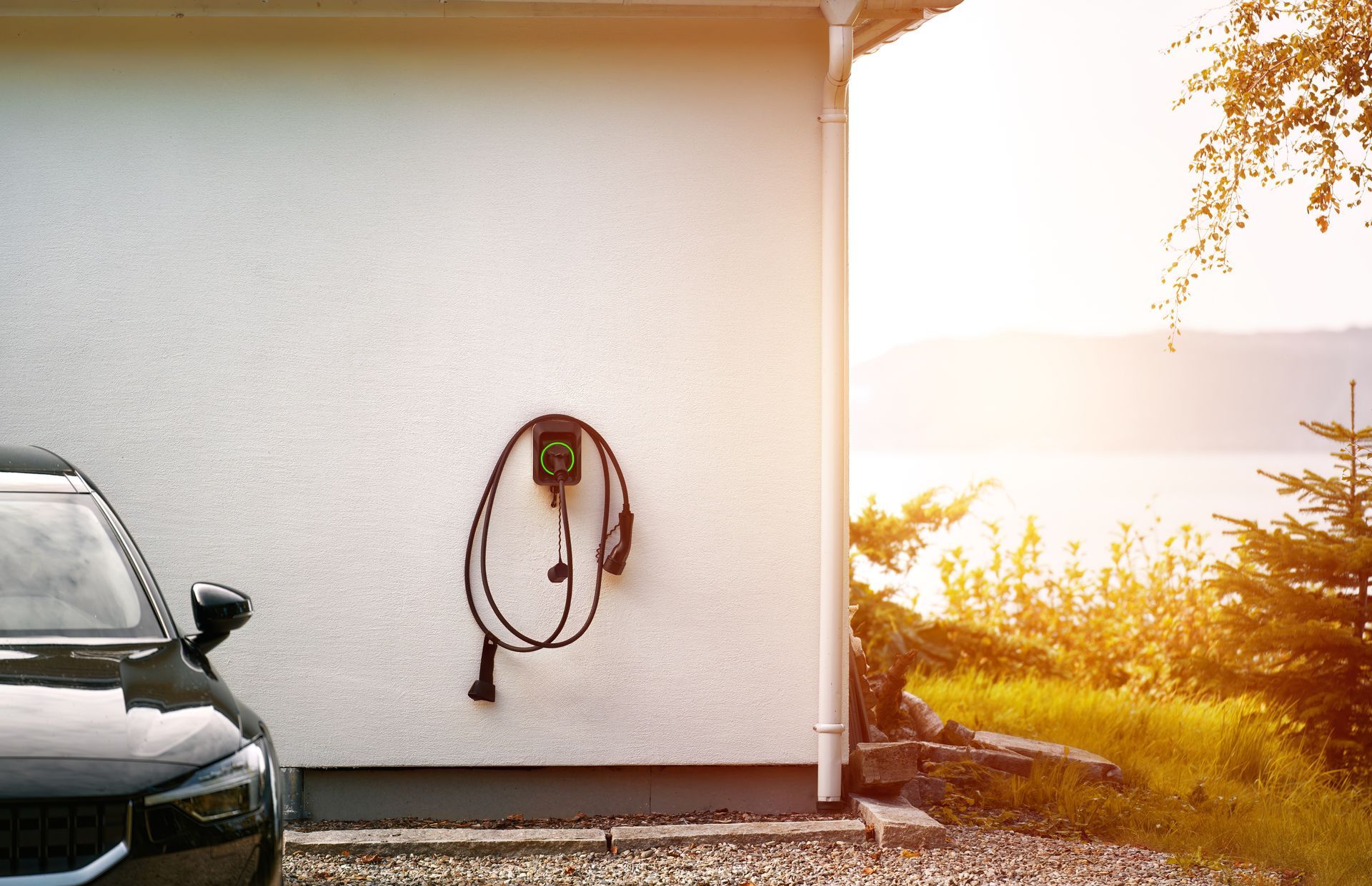 Black car beside a white house with a hose reel, overlooking a bright sunset view
