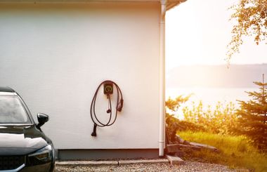 Black car beside a white house with a hose reel, overlooking a bright sunset view