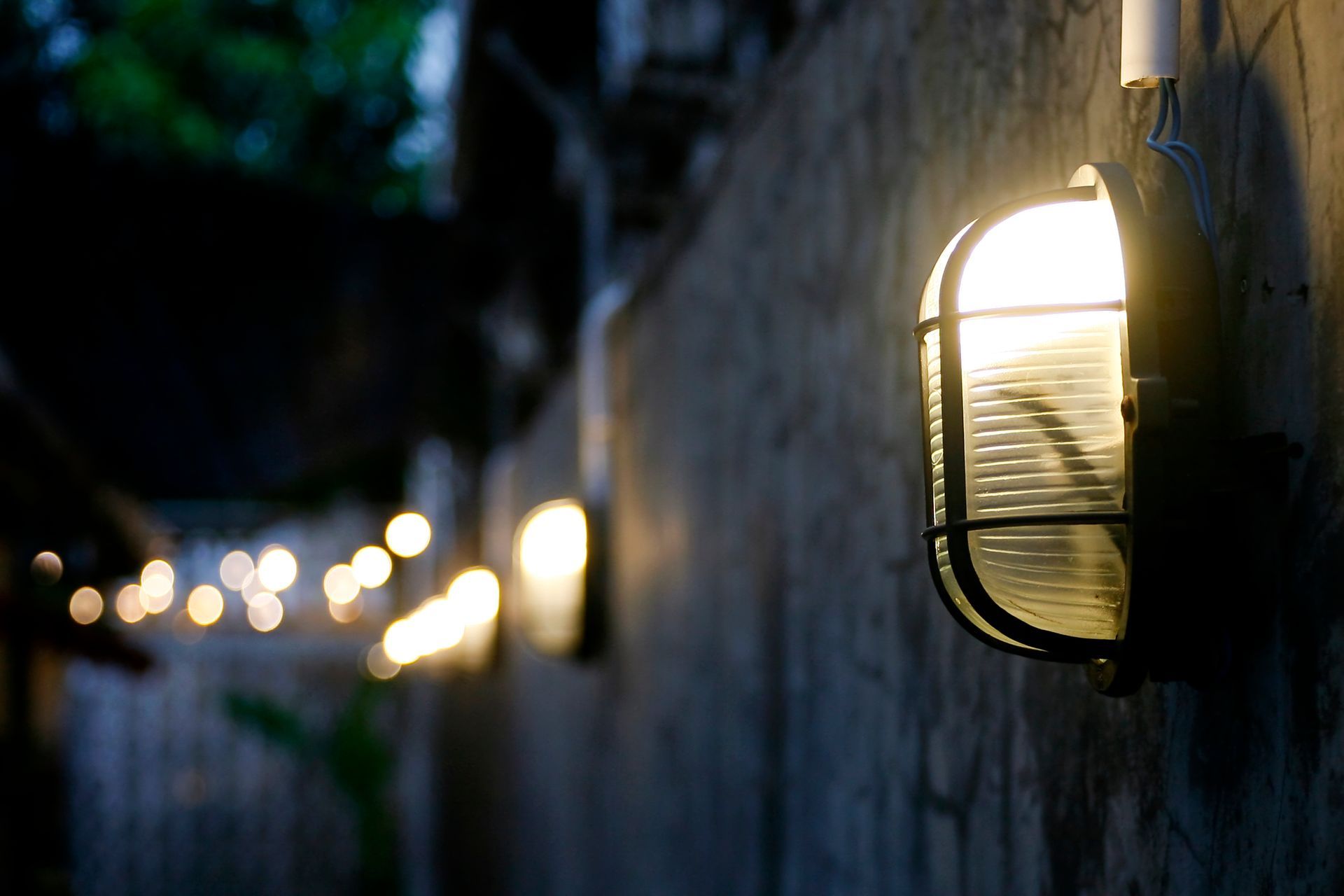 Dim wall lantern glowing on a stone wall at dusk, with a row of blurred lights receding into the background