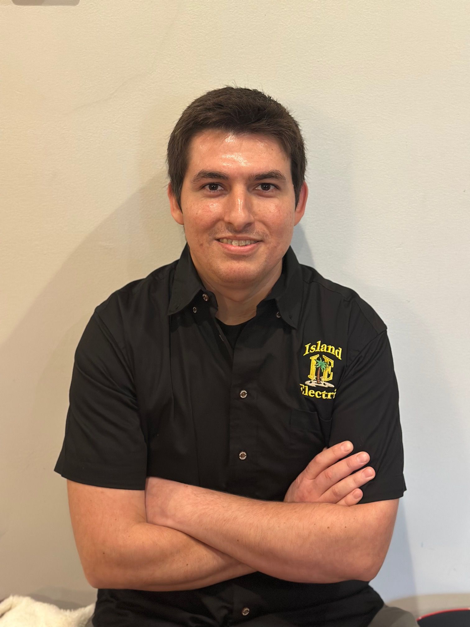 Man standing with arms crossed, wearing a black collared shirt, against a plain beige wall.
