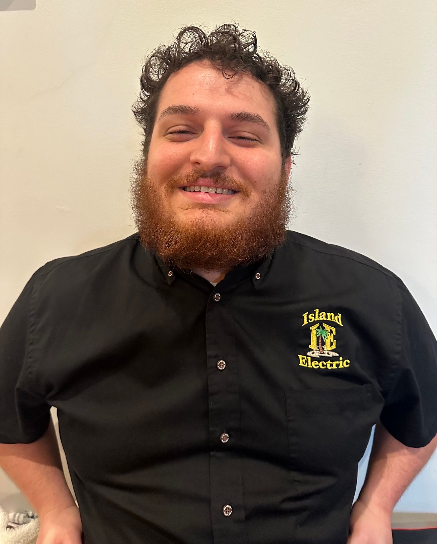 Smiling person in a black polo shirt with a yellow logo, standing against a plain wall.