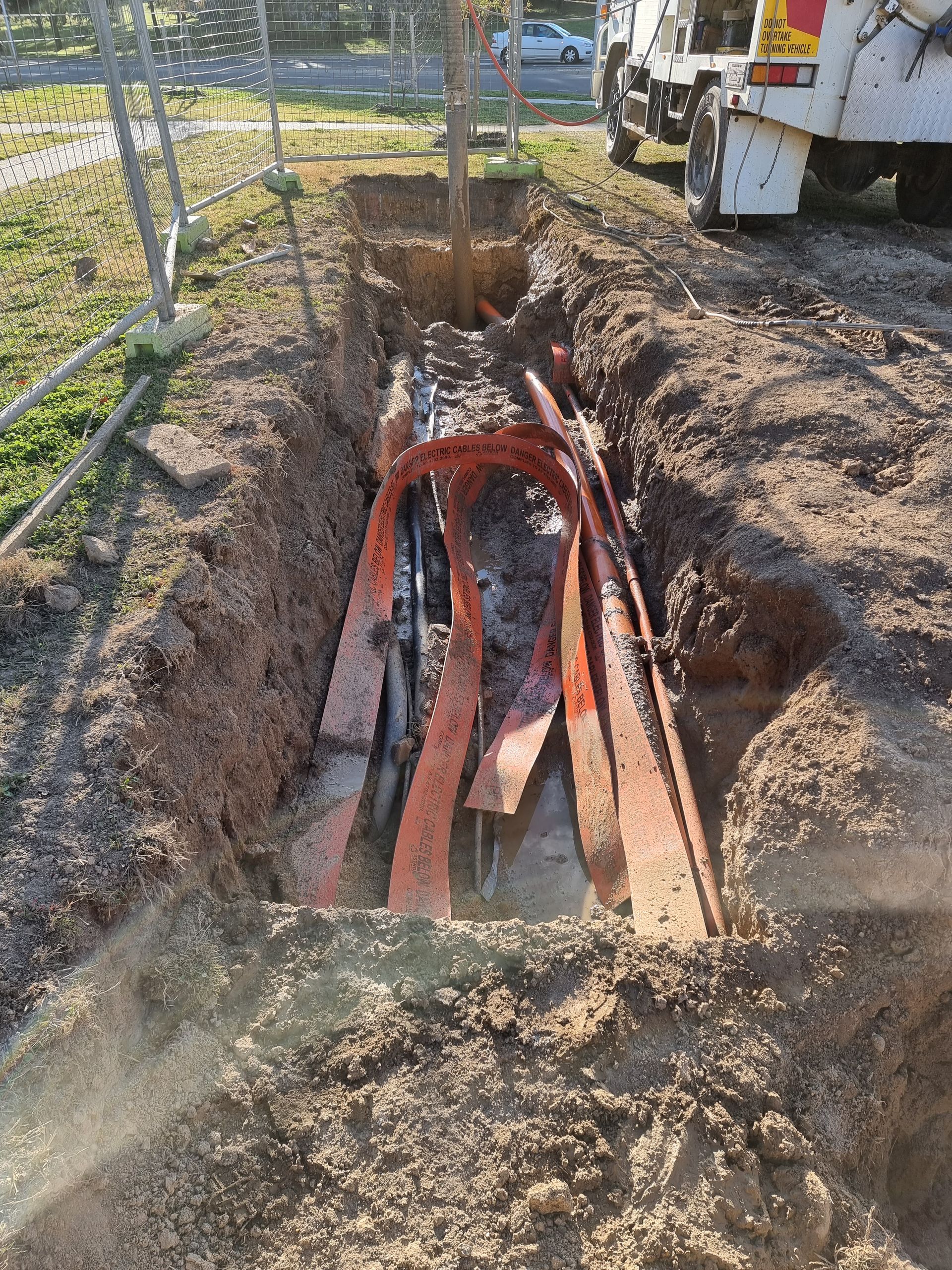 Trench with exposed orange cables and conduits; construction site with truck and fence — Pro Pipe Solutions in Queanbeyan, NSW