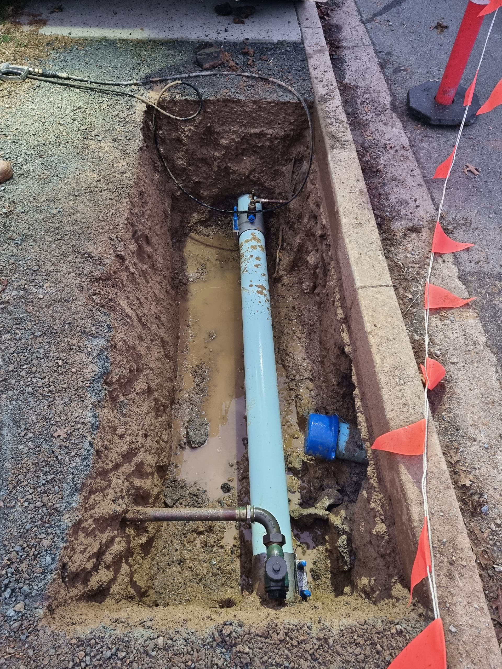 Open trench with a large blue pipe, possibly water main, in the ground near a curb. Orange flags are nearby — Pro Pipe Solutions in Queanbeyan, NSW