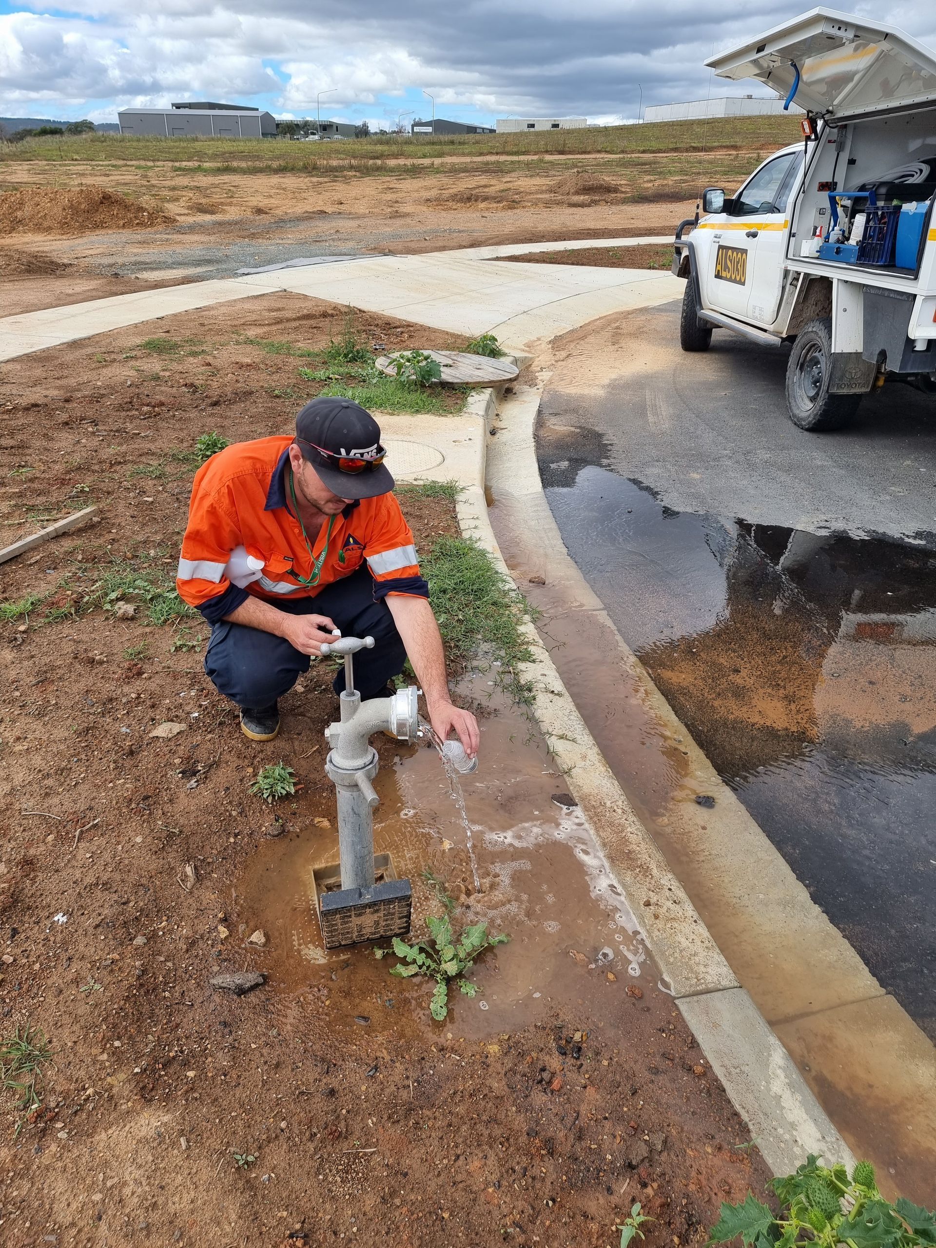 Man in work clothes testing water from a faucet, standing next to a vehicle and puddle — Pro Pipe Solutions in Queanbeyan, NSW