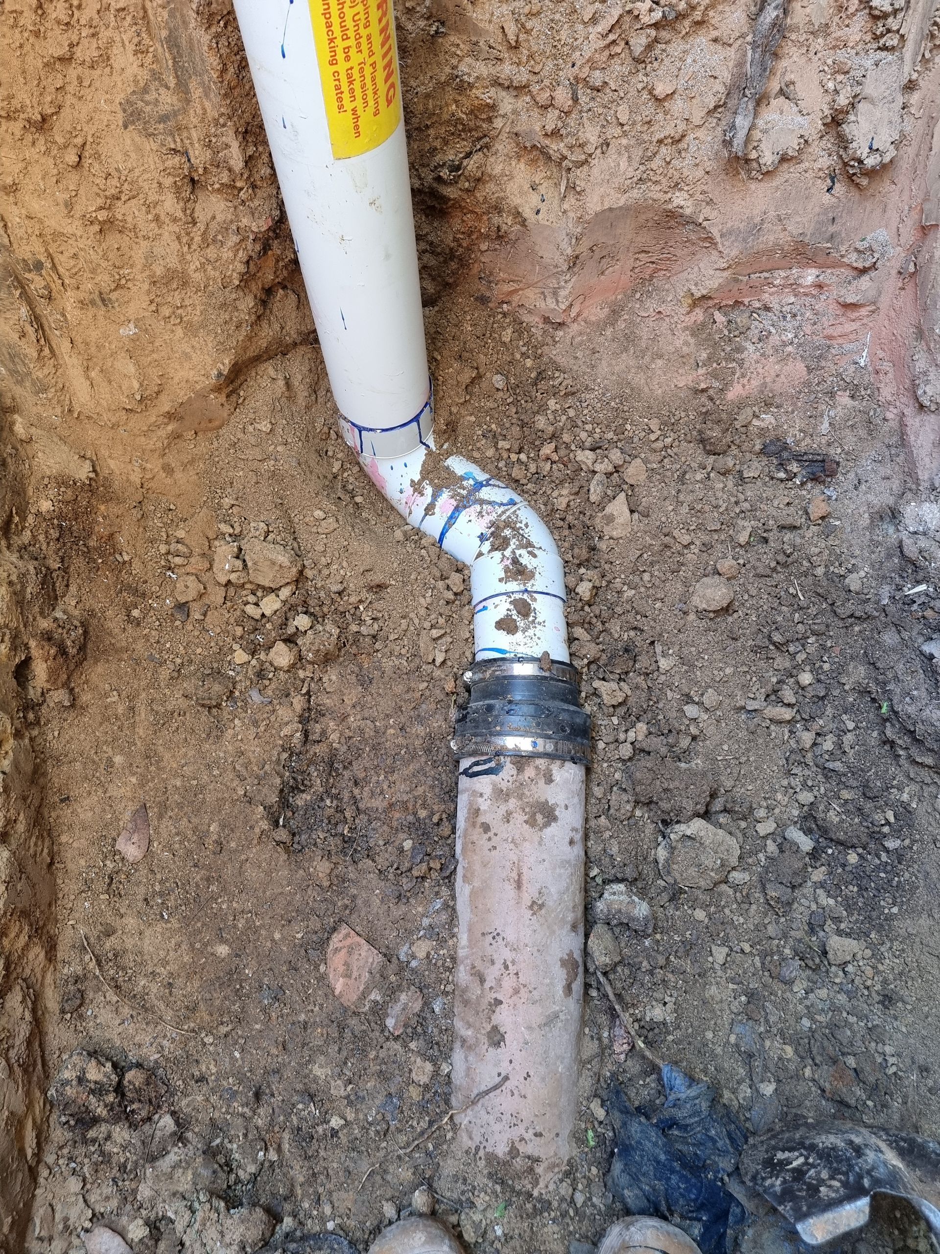 White PVC pipe connected to a clay pipe in a dirt trench — Pro Pipe Solutions in Queanbeyan, NSW