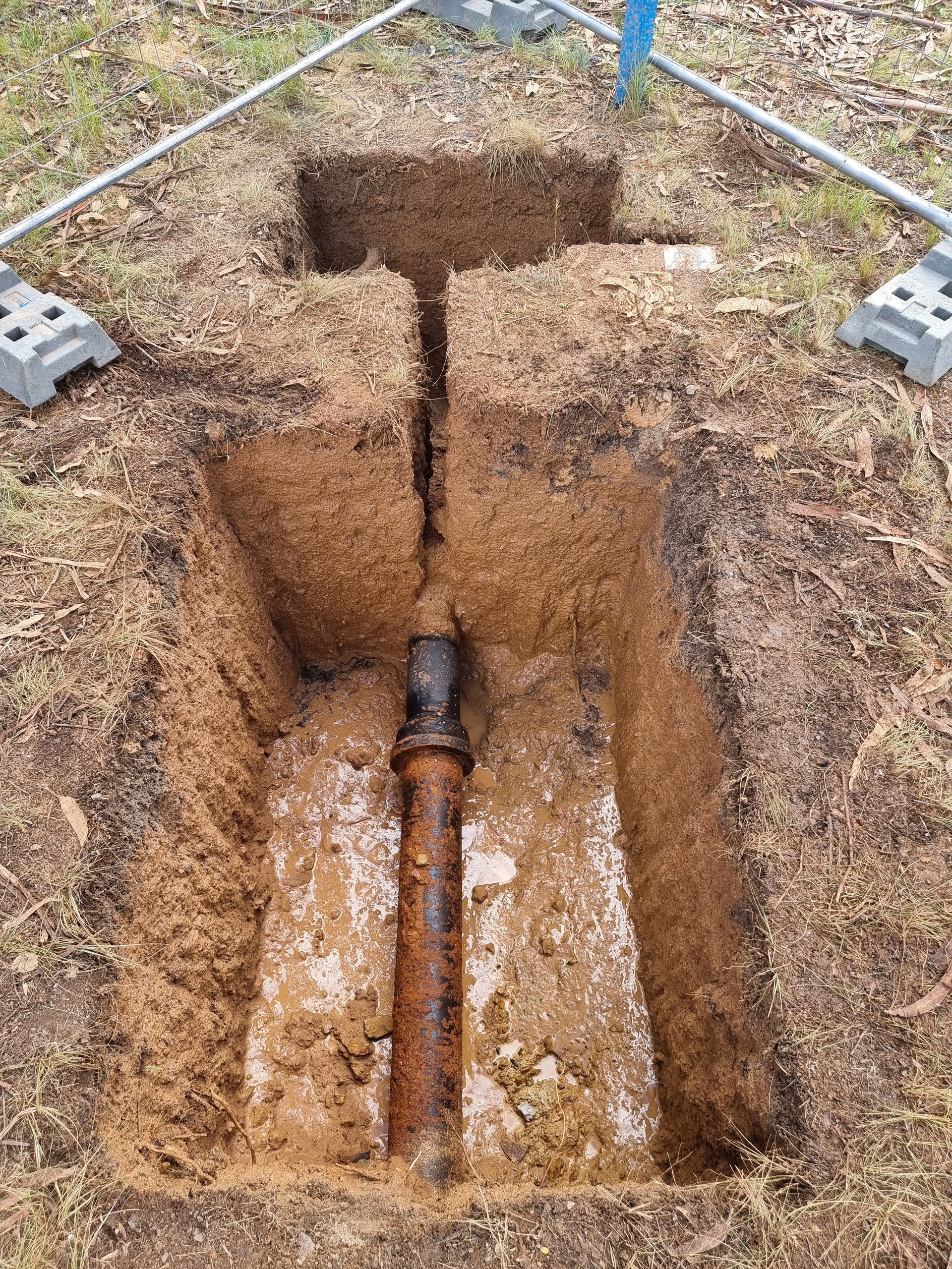 Trench in grass revealing a rusty pipe. Ground is dug up around the pipe, with supports visible at the edges — Pro Pipe Solutions in Queanbeyan, NSW