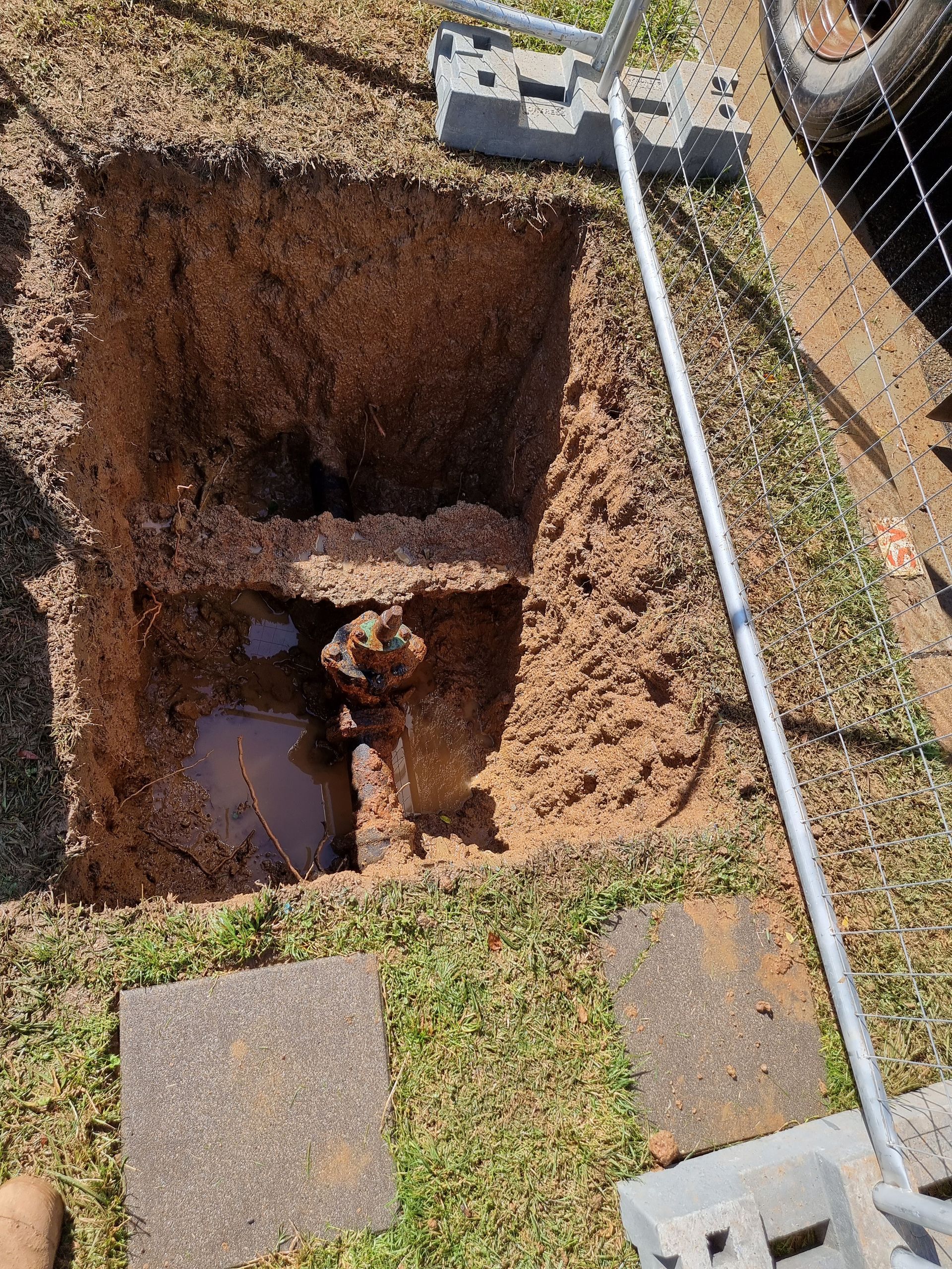 An open hole in the ground reveals buried pipes and fittings, with water pooling at the bottom and a metal fence nearby — Pro Pipe Solutions in Queanbeyan, NSW