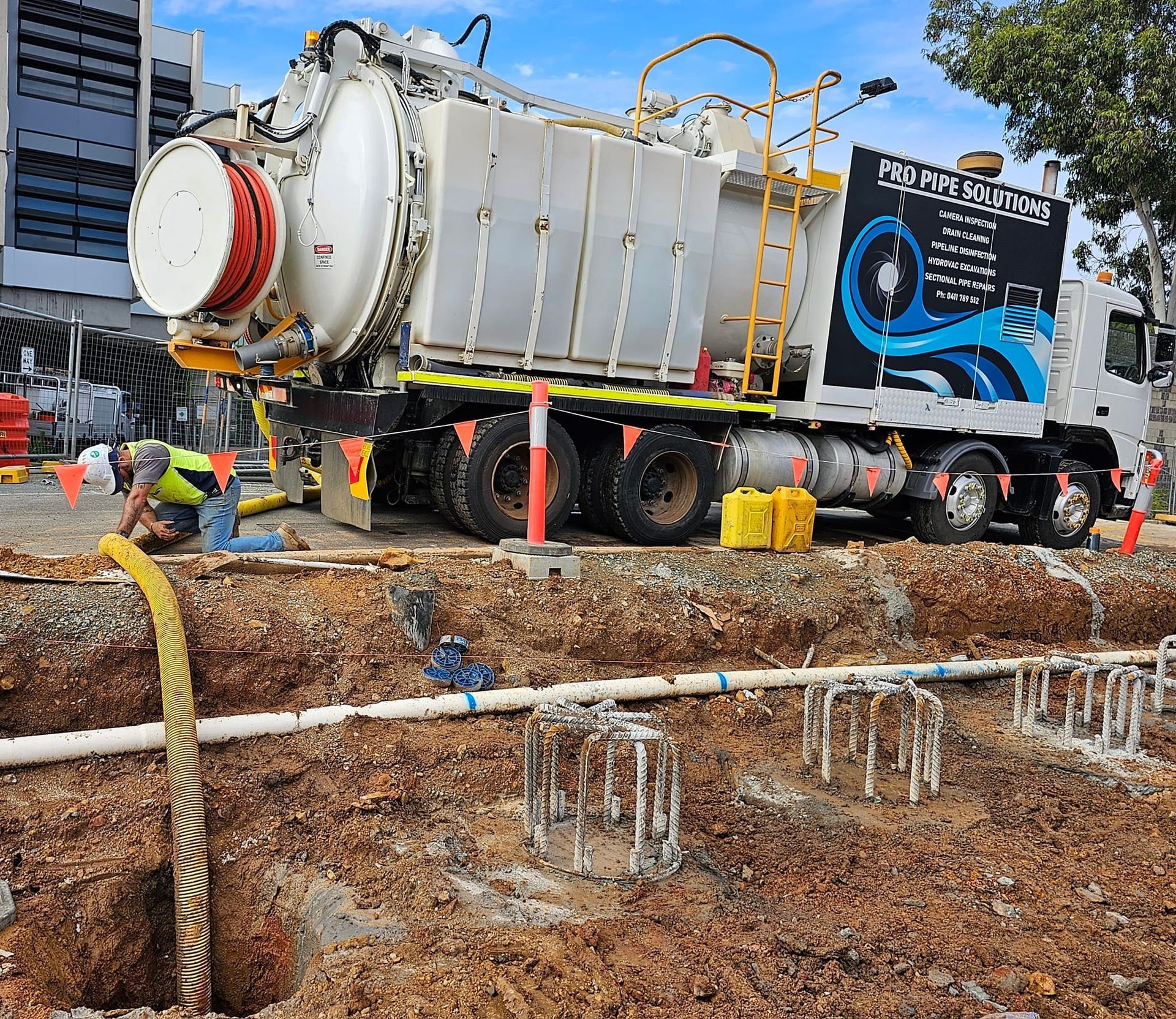 Hydro-excavation truck at construction site. Worker operates hose over open pit. Cloudy sky, building visible  — Pro Pipe Solutions in Queanbeyan, NSW