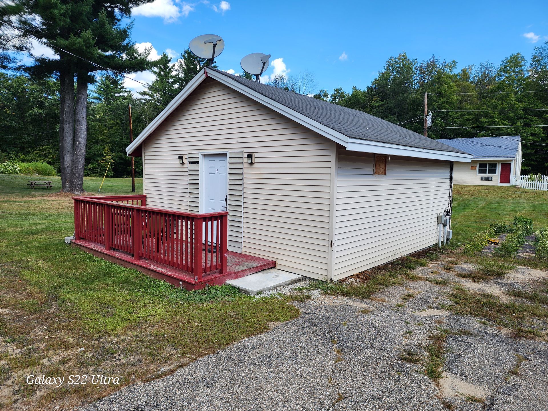 Small white utility building with red porch railing and satellite dishes, beside a gravel path and trees.