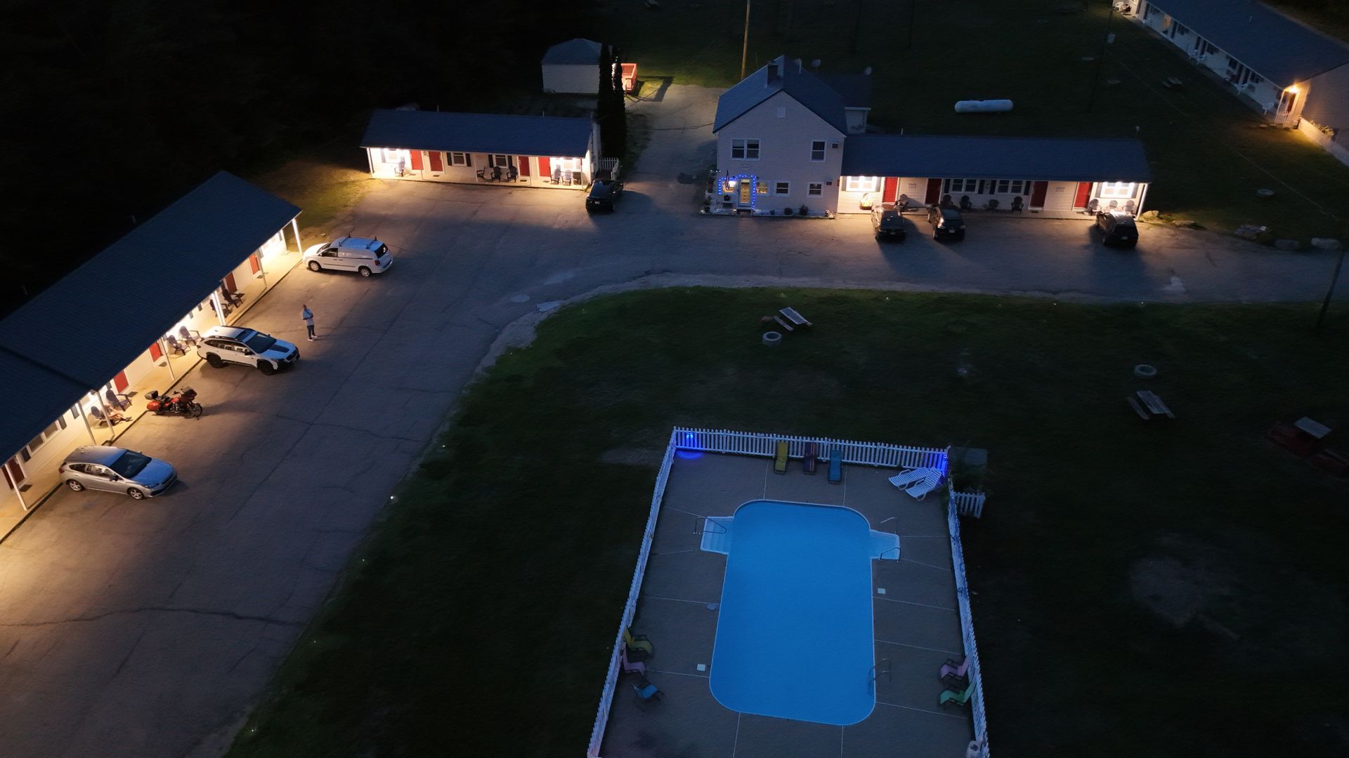 Aerial view of a lit outdoor pool at night beside a building and parking area