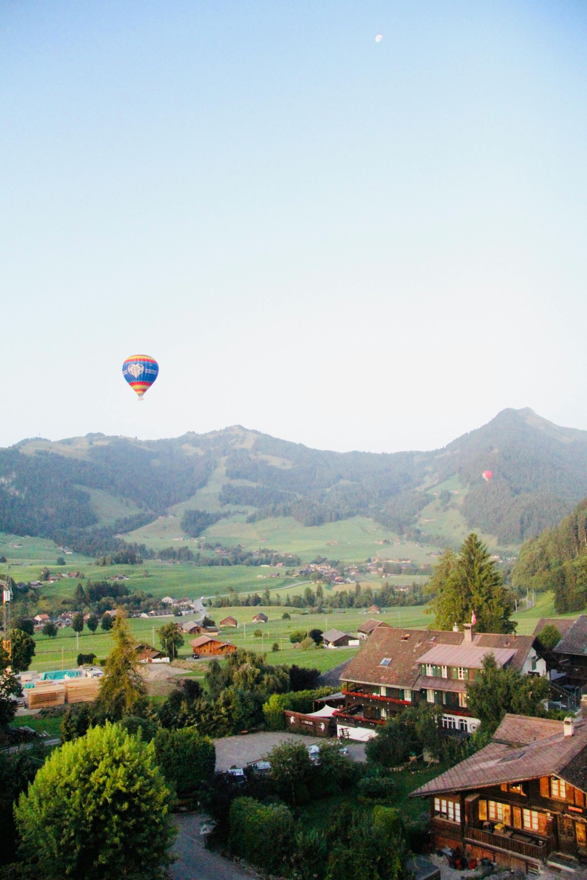 Hot air balloon floating over a green valley with hills, village houses, and a clear blue sky at sunrise.
