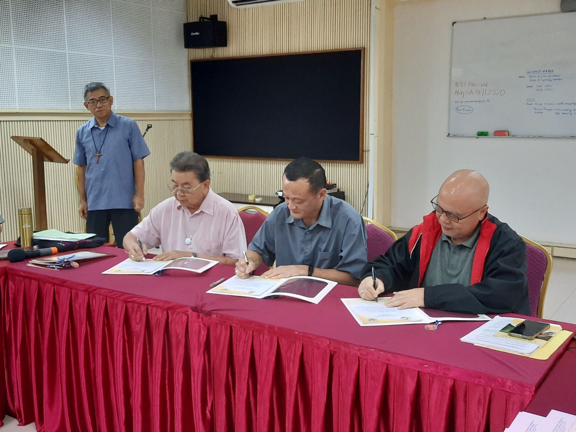 Four men signing documents at a table covered with a red cloth; one man observes from behind a podium.