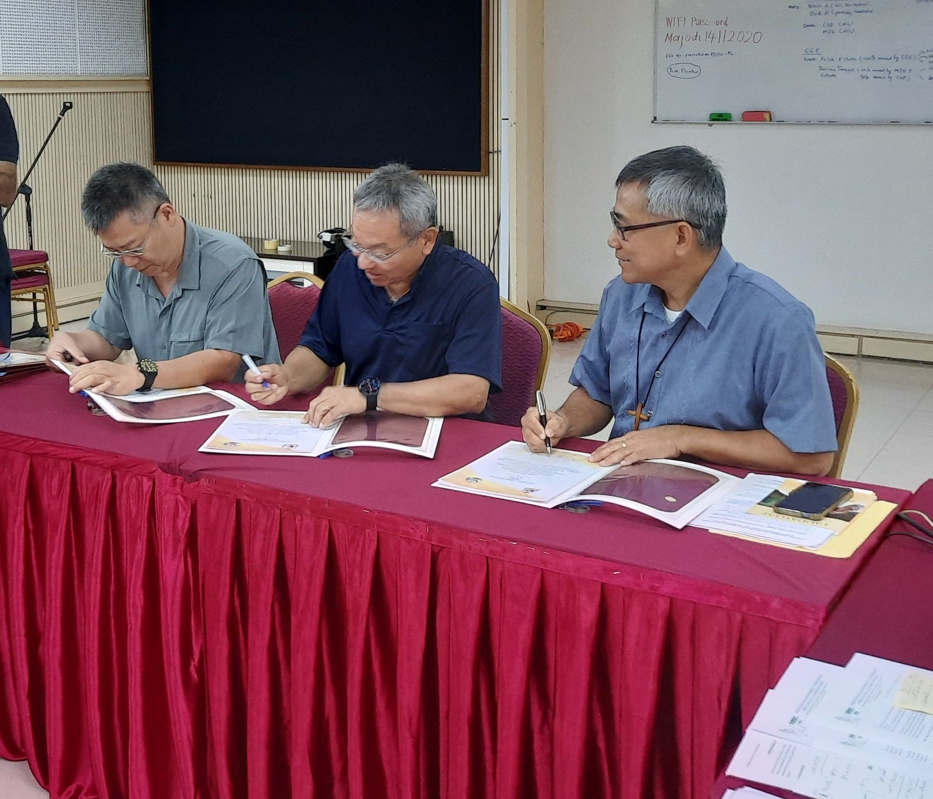 Three men signing documents at a red table in a room.