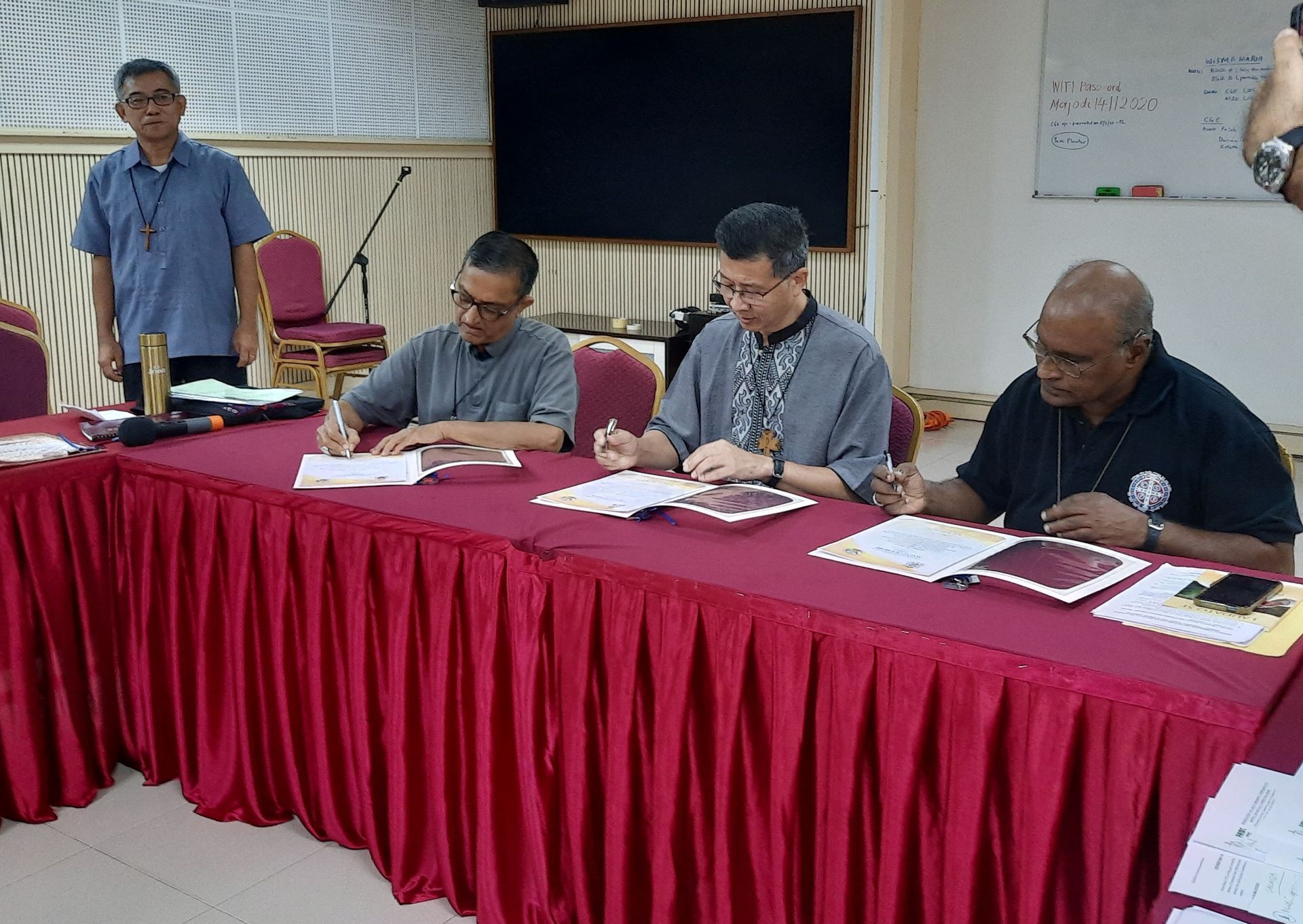 Four people sign documents at a table covered in a red cloth. One man stands behind them.
