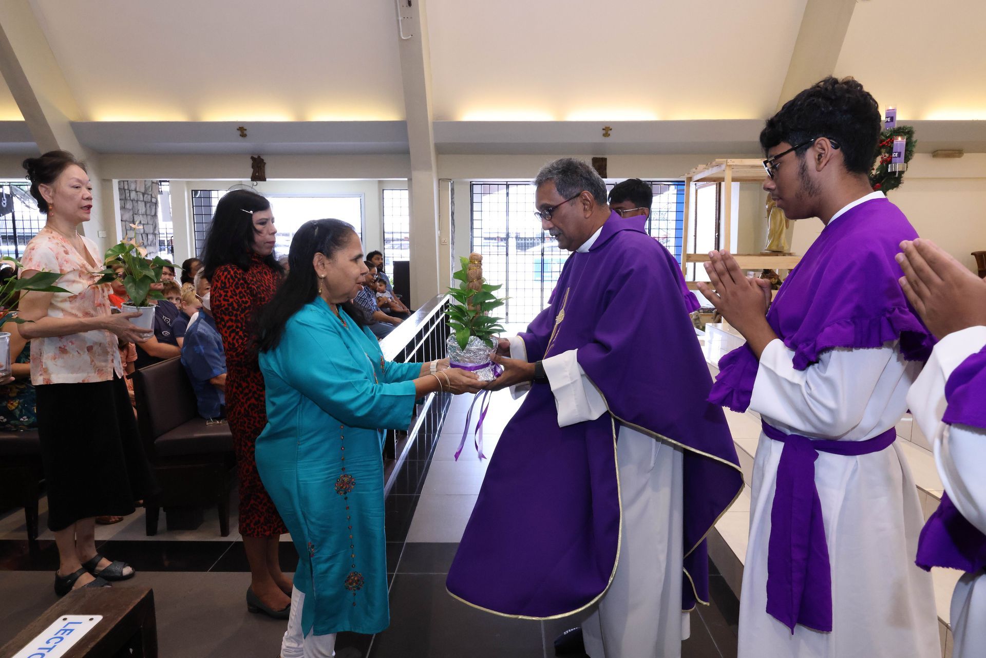 A priest in purple robes receiving a gift from a woman in a turquoise outfit inside a church.