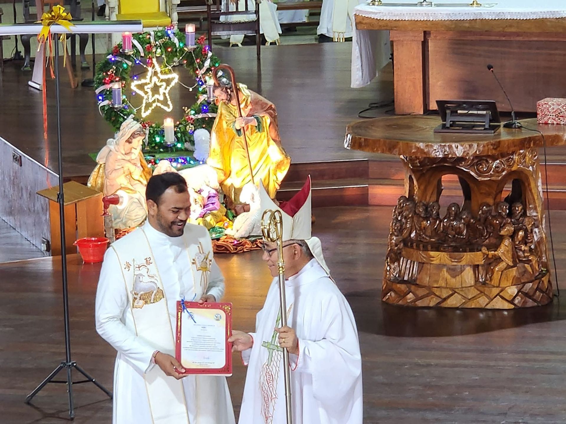 Two clergy in white robes, one presenting a document, with a nativity scene in the background.