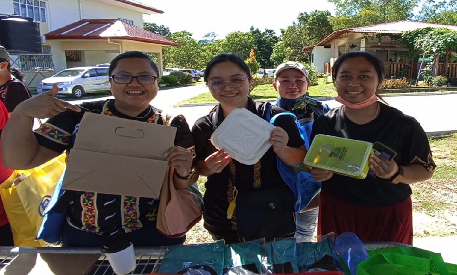 Three people smiling, holding cardboard and containers, outdoors near a table and buildings.